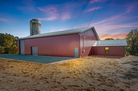 Red post-frame pole barn at dusk beside a farm silo, under a violet sky